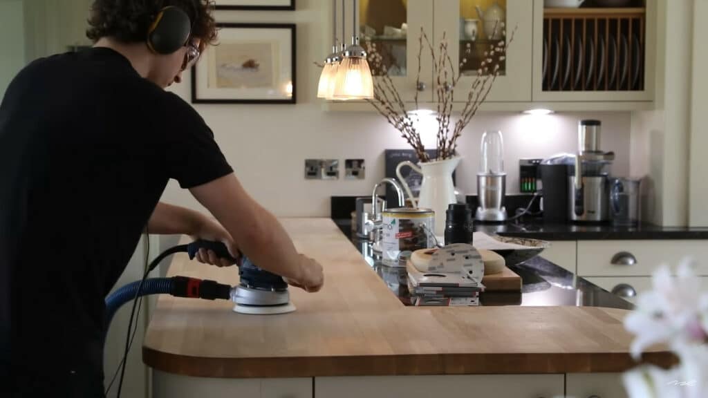 A person using an orbital sander on a wooden countertop in a well-equipped kitchen.