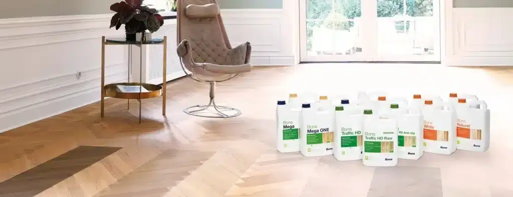 A luxurious living room with herringbone wooden flooring is featured, showcasing a variety of Bona wood floor finish products placed prominently on the floor. The finishes include options like Bona Mega, Bona Traffic HD, and others, providing a range of choices for wood floor treatment. A stylish chair and a small table with a plant add to the elegant decor of the room, which is brightly lit by natural light from large glass doors.