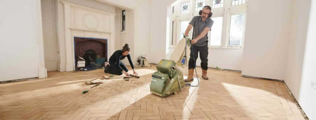 A professional from Mr Sander® sands a floor in a living room in, UK using the Lagler HUMMEL® floor sanding machine. The HUMMEL® features a powerful LED lighting system, 2.2 kW single-phase AC motor, 2400 rpm sanding drum speed, 200 mm drum width, and 200 x 750 mm sanding belt. The machine has extremely low dust emissions, a weight of 79 kg, and is easy to maintain and service.