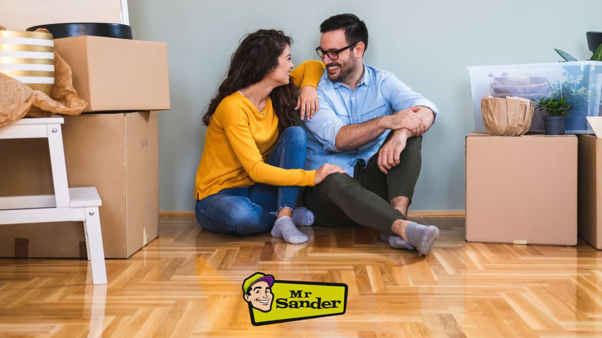 A happy couple sits on a beautifully restored parquet floor, completed by Mr Sander® in Shepherds Bush. Surrounded by moving boxes, they smile at each other, enjoying their newly refinished home.