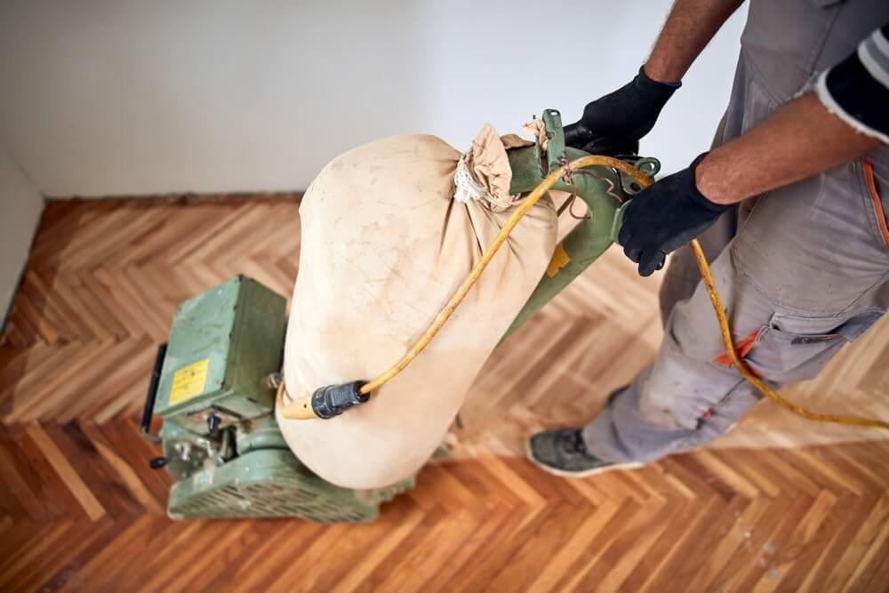 A Mr Sander® technician sanding a parquet floor in a Upminster living room using a LAGLER HUMMEL® machine. Features include powerful LED lighting, high powered aggressive sanding, extremely low dust emissions, quick belt changes, 30+ years lifespan, and easy maintenance. Technical specs: 230 V / 50 CPS / 2.2 kW motor, 200 mm drum width, 2400 rpm drum speed, 200 x 750 mm belt dimensions, 79 kg weight.