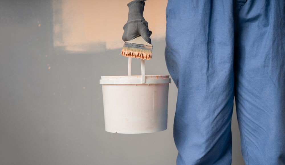 A professional painter holding a bucket and brush, wearing blue work trousers, illustrating the dilemma: “Should I Paint Before I Sand the Floors?”