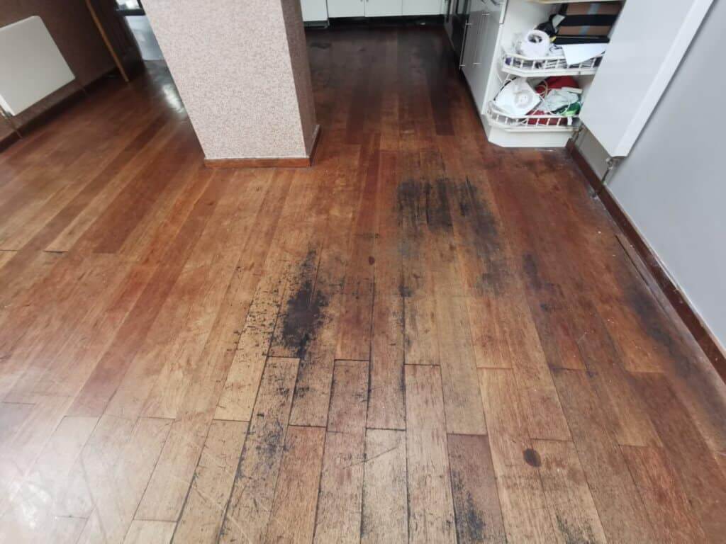 A heavily worn and stained hardwood floor with visible dark patches and scratches, indicating the need for refinishing. The floor shows signs of heavy traffic, with large black marks in high-use areas near the kitchen. The wood appears dull and damaged, with the finish completely worn away in places, making it a prime candidate for sanding and restoration.
