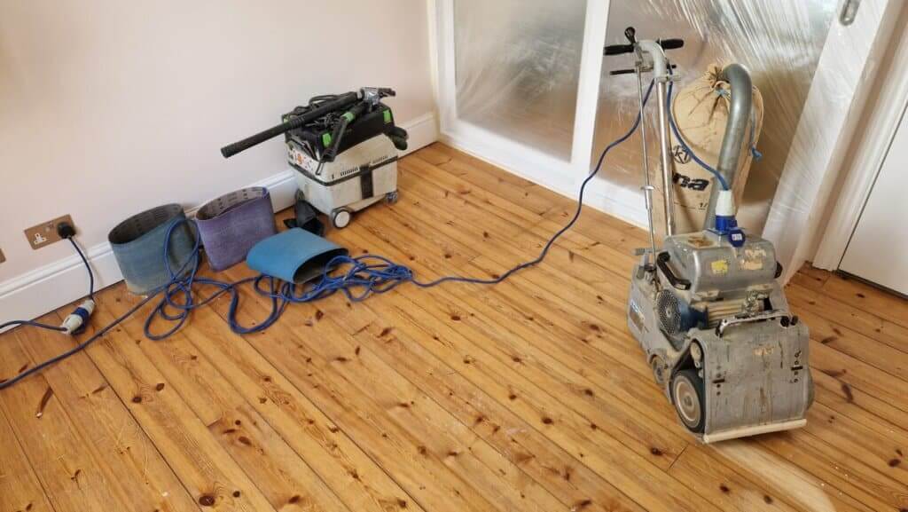 A man from Mr Sander® is sanding a wooden floor in living room using a Bona Belt professional belt sander (Model AMO110000.3). The machine has a drum width of 200 & 250 mm, uses sanding belts sized 200 x 750 mm or 250 x 750 mm, and has a motor power of 2.2 kW. The sander weighs 74 kg.