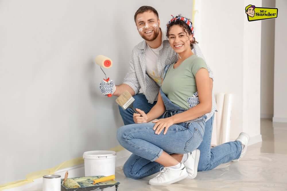 A happy couple kneeling while painting a wall light grey. They hold a paint roller and brush, with painting supplies on the floor, showcasing teamwork during a home renovation.