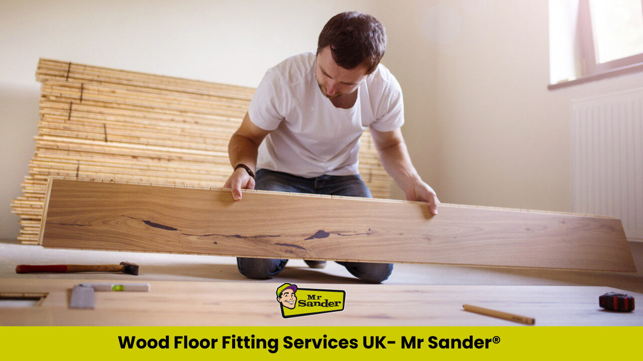 A man wearing a white T-shirt carefully positioning a solid oak wood plank during a wood floor fitting project in a bright, partially renovated room.