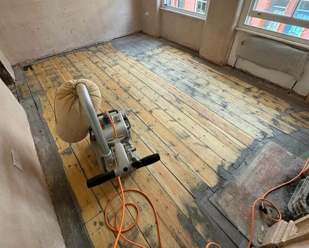 A professional from Mr Sander® is sanding a pine wood floor in a living room in ,UK using a Bona Belt sander (AMO110000.3). The machine features a 2.2 kW motor, 230 V voltage, 50 Hz frequency, and a sanding belt size of 200 x 750 mm or 250 x 750 mm. The total weight is 74 kg.