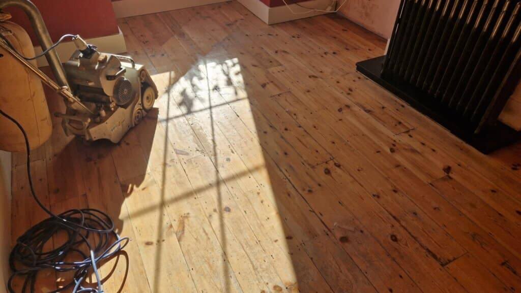 A professional from Mr Sander® is sanding a pine floor in South Woodford, UK living room using a Bona Belt sander (AMO110000.3). The machine features a 2.2 kW motor, 230 V voltage, 50 Hz frequency, and a sanding belt size of 200 x 750 mm or 250 x 750 mm. The total weight is 74 kg.