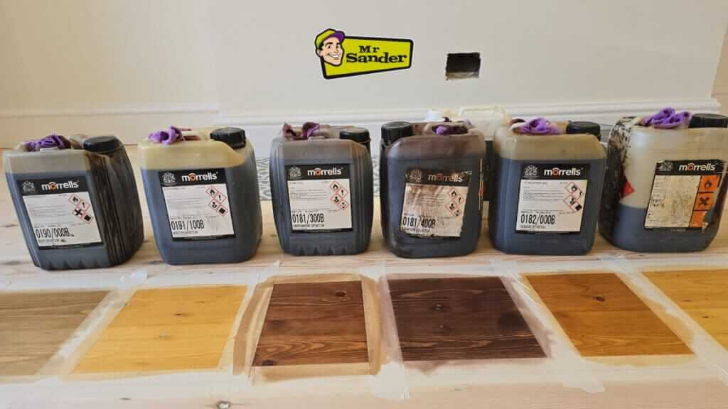 A variety of Morrells wood stains and finishes displayed alongside wood samples showcasing different stain shades on a wooden floor.