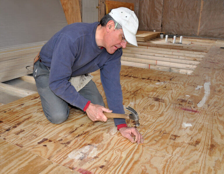 A floor installer kneeling and hammering nails into a plywood subfloor, demonstrating proper preparation for West London Floor Fitting.