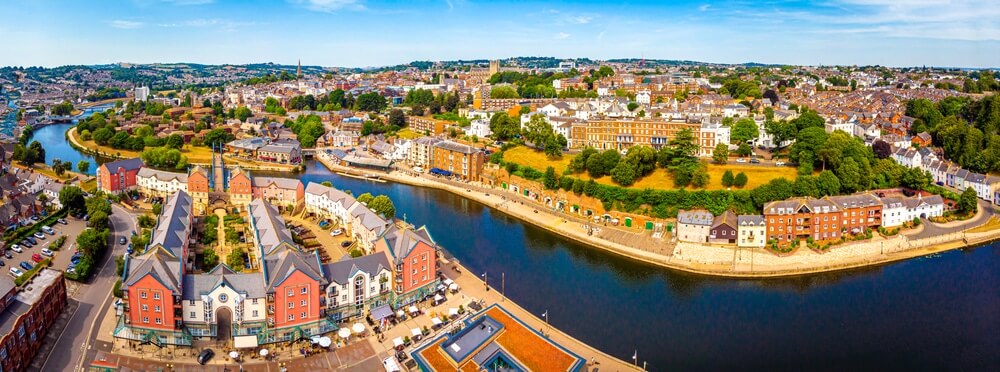 A panoramic aerial view of a riverside town in the UK on a bright summer day, featuring colorful waterside buildings and lush greenery under a clear blue sky.