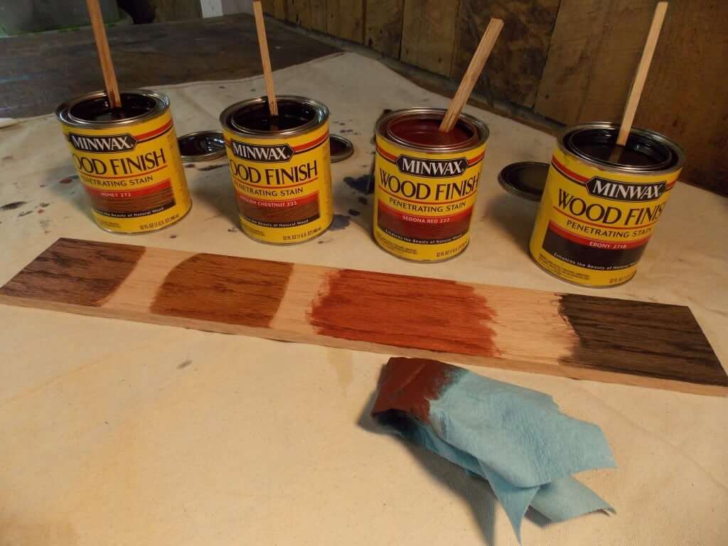 Four cans of Minwax Wood Finish penetrating stain, each with a stir stick, are displayed on a table. In front of the cans, a piece of wood shows various stain colors being tested, including dark brown, honey, reddish, and ebony shades. A blue cloth with some stain residue is also visible on the table.