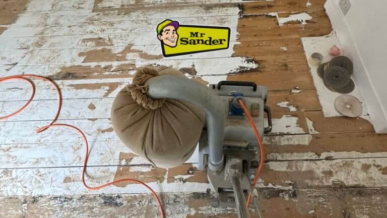 A large floor sander is being used to strip away multiple layers of old paint from a wooden floor. The floor is made of white pine and is in rough condition.