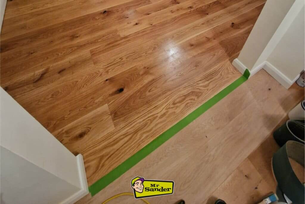 A freshly finished wooden floor in a living room with a fireplace, mirror, and chandelier, showcasing the result of applying floor finish in sections.