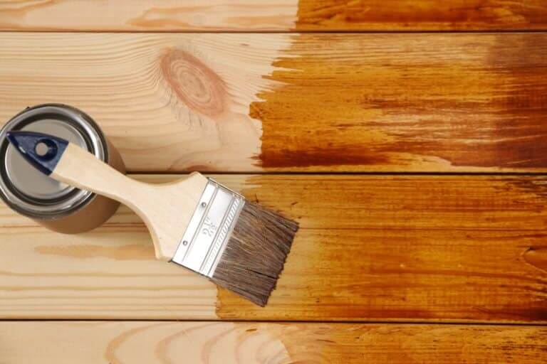 A paintbrush and a can of varnish placed on a wooden floor. The floor shows a clear distinction between the unfinished light wood and the darker, shiny wood that has been refinished with varnish. This image illustrates the process of floor sanding and refinishing.