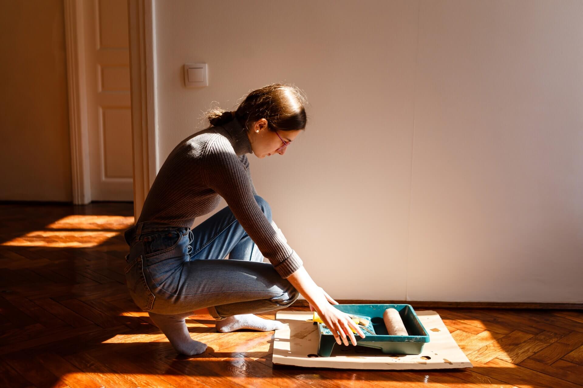 A woman kneeling on a wooden floor, carefully preparing finishing materials in a paint tray, illustrating the effort involved in DIY floor refinishing.