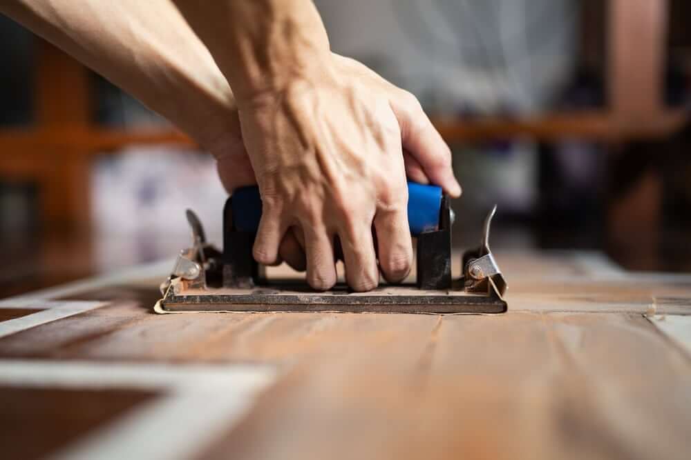 A close-up view of a person using a handheld floor sander on a wooden floor.