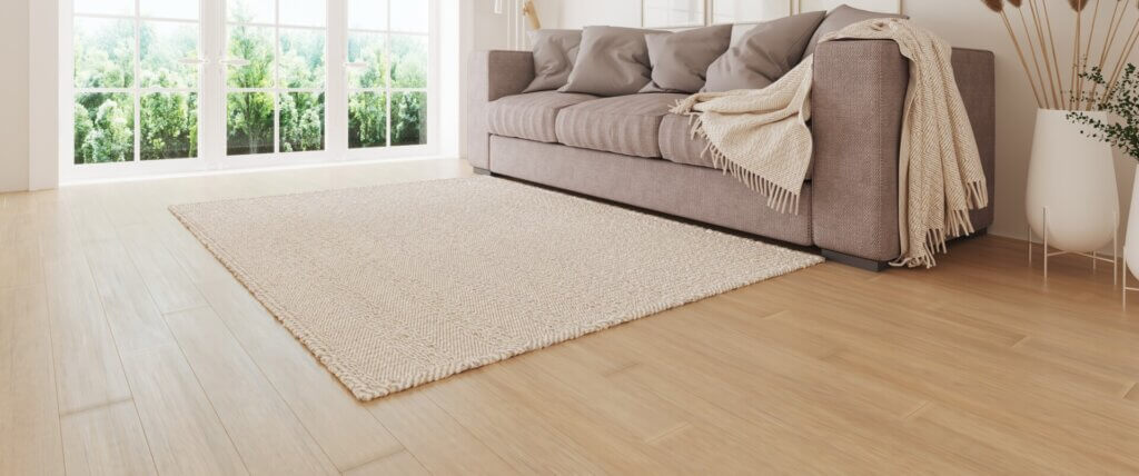 Bright living room with large windows and a comfortable grey sofa, featuring a soft beige rug on newly refinished wooden floors by Mr Sander®. The natural light enhances the smooth, polished finish of the light wood flooring.