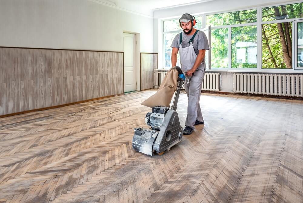 A professional floor refinisher wearing ear protectors sands a large herringbone floor in a bright, open venue, illustrating the importance of careful planning and adequate drying times for theatrical or gallery spaces.