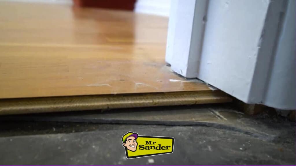 A close-up image of a damaged laminate floor. The floor is peeling away from the wall, revealing the underlayment and subfloor.