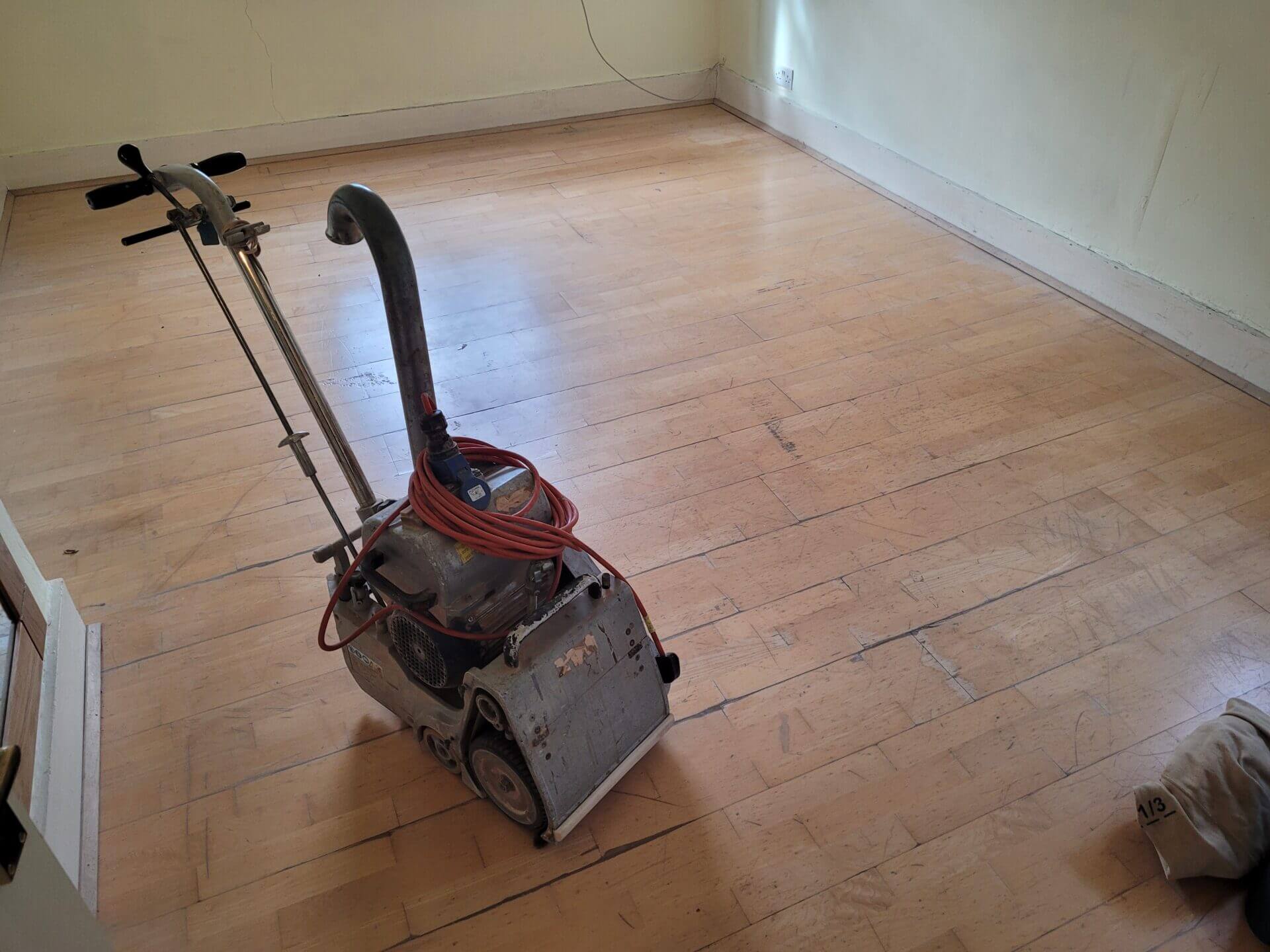 A heavily worn maple floor in a living area with a professional floor sanding machine at the centre, highlighting the need for floor sanding for maple floors.