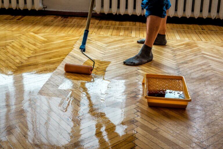 A technician applying fresh lacquer with a roller to a partially refinished parquet floor, illustrating how to transform tired floors.