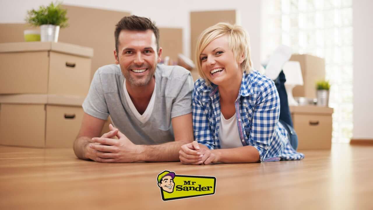 Happy couple enjoying their newly restored wooden floor by Mr Sander, surrounded by moving boxes in a bright and modern home.