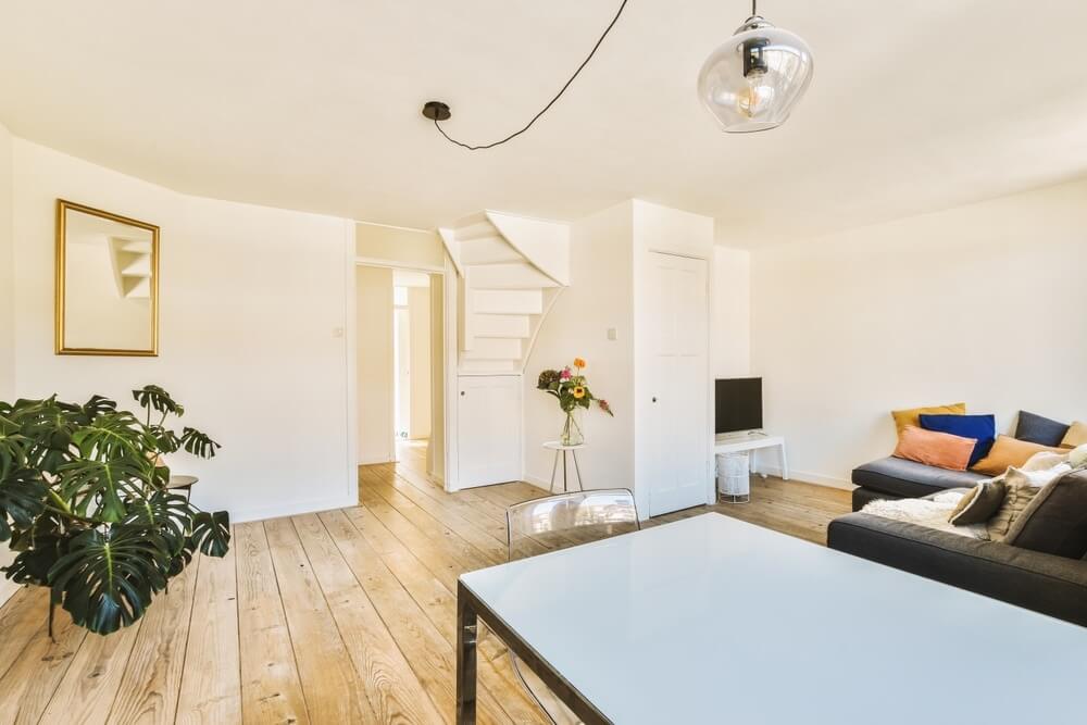A modern living room in Enfield, UK, showcasing beautifully sanded and refinished light oak hardwood floors. The room is bright and minimalist, featuring a comfortable gray sofa with colorful cushions, a white table, a potted plant, and a spiral staircase leading to the upper floor.