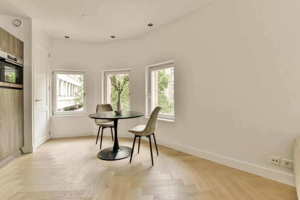 A modern dining area in Richmond, UK, featuring a beautifully sanded and refinished light oak herringbone parquet floor. The room is furnished with a round black table and two beige chairs, with three windows providing ample natural light.