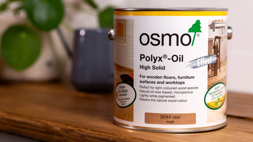 A can of Osmo Polyx-Oil for wood finishing on a wooden countertop with blurred plant and teapot in the background.