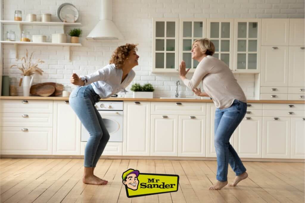 Two women happily dancing barefoot on a beautifully refinished light oak wood floor in a modern kitchen. The kitchen features white cabinetry, open shelving, and minimalist decor, creating a bright and airy atmosphere. The light oak floor adds warmth and complements the clean, contemporary design of the space. The Mr. Sander® logo is visible at the bottom of the image.
