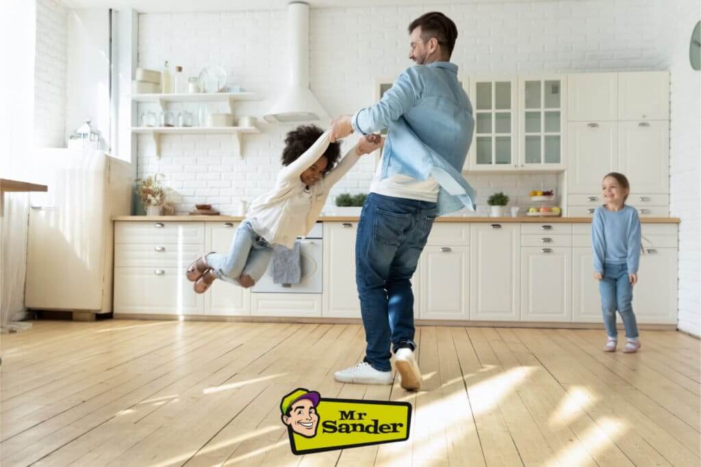 A joyful family moment captured in a bright kitchen with a beautifully refinished light pine wood floor. A father spins his young daughter around while another child watches and smiles. The kitchen features white cabinetry, open shelving, and a minimalist design, complementing the warm tones of the polished pine floor. Sunlight streams in, reflecting off the smooth floor surface, adding a lively and warm atmosphere. The Mr. Sander® logo is displayed at the bottom center.