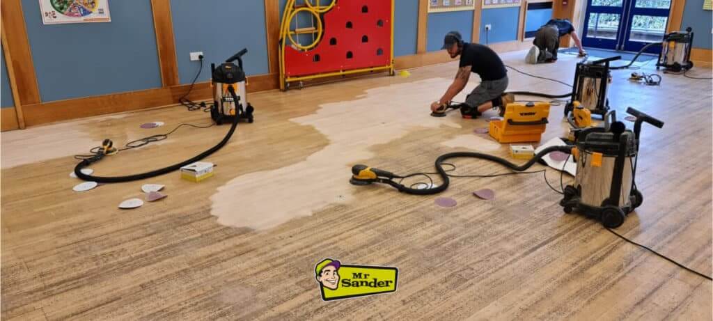 Professionals sanding and refinishing a large oak wood floor in a community or school hall using industrial-grade equipment, part of a project by Mr Sander®