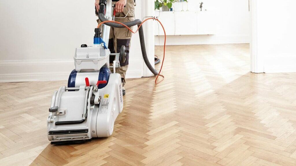 A man from Mr Sander® sanding a herringbone parquet floor in a living room in Hammersmith, UK using the Bona Belt UX (AMK173100). The sander has a motor power of 2.2 kW, voltage of 230 V, frequency of 50 Hz, and a sanding belt size of 200x750 mm. It also features a 200 mm drum width and weighs 85 kg, providing efficient and precise sanding.