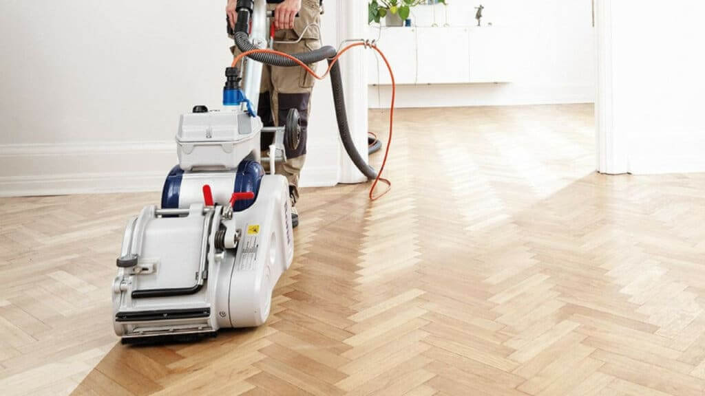 A man from Mr Sander® sanding a herringbone parquet floor in a living room in Holloway, UK using the Bona Belt UX (AMK173100). The sander has a motor power of 2.2 kW, voltage of 230 V, frequency of 50 Hz, and a sanding belt size of 200x750 mm. It also features a 200 mm drum width and weighs 85 kg, providing efficient and precise sanding.