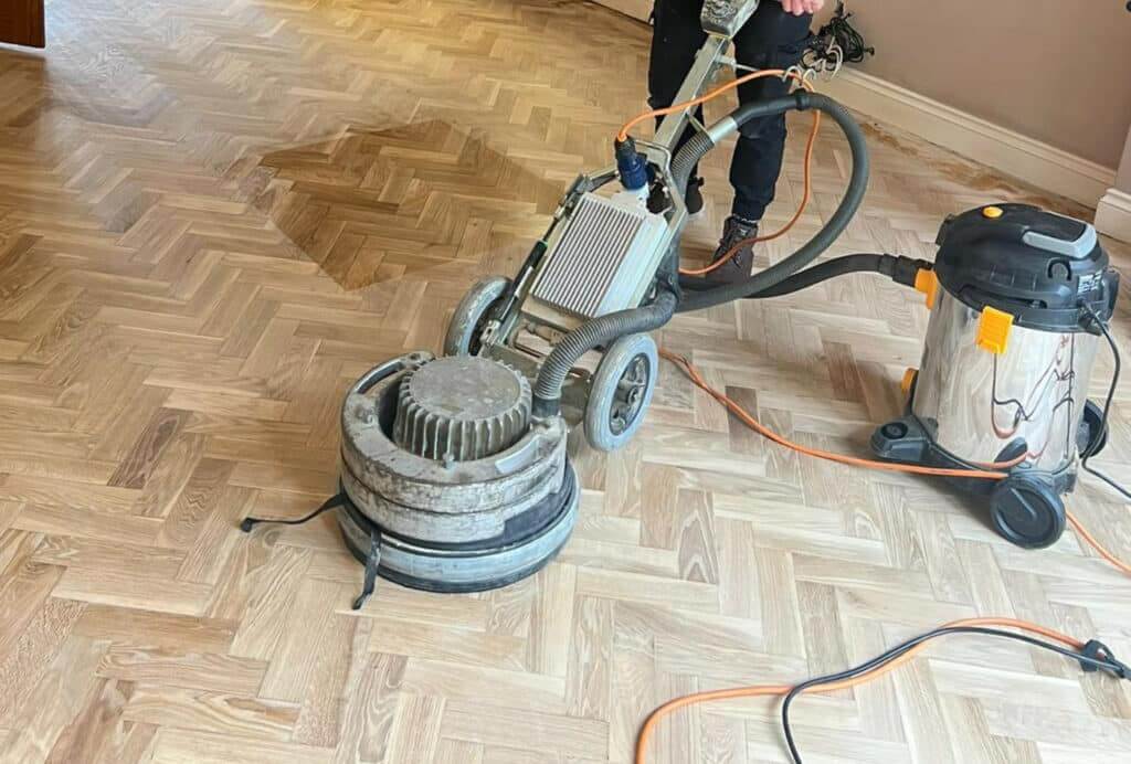 A man from Mr Sander® sanding a parquet floor in a living room in Beddington, UK, using the PALLMANN SPIDER wood floor sanding machine. The PALLMANN SPIDER features 360° illumination, switchable power reserve, triple-disc technology, infinite speed adjustment (50-400 RPM), a 230 V power supply, 2000 W power consumption, 400 mm sanding width, 42 kg weight, and an additional weight of 15 kg for increased sanding pressure.