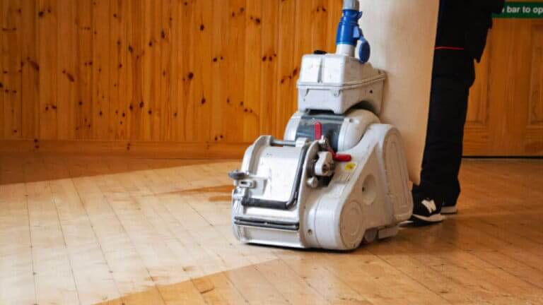 A professional-grade floor sanding machine in action on a light-colored wooden floor, with a wooden-paneled wall in the background.