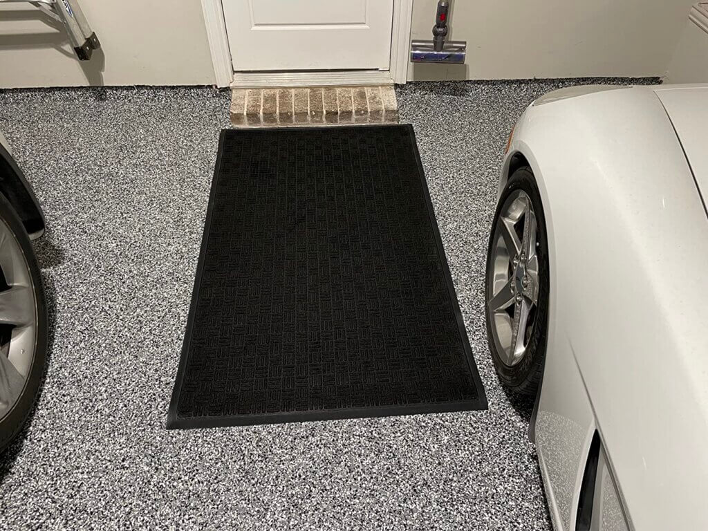 A garage interior with a speckled epoxy-coated floor, featuring a black mat placed near a door with a small brick threshold. A white car is parked on the right, and part of another vehicle is visible on the left. The durable and polished floor showcases professional finishing.