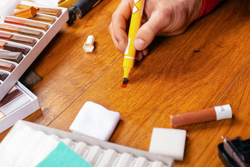 Close-up of a person using a wood repair marker to fill a scratch on a wooden floor, showcasing DIY solutions for scratched wooden floors.