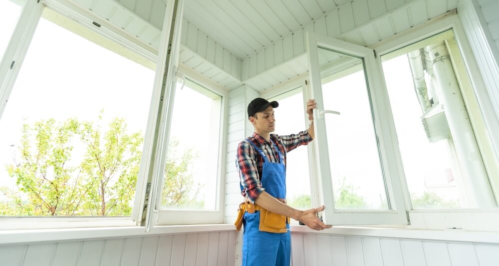 A professional wearing a checkered shirt, blue overalls, and a black cap inspects an open window in a bright, white-paneled room. The floor appears freshly sanded and refinished, complementing the clean and modern aesthetic.
