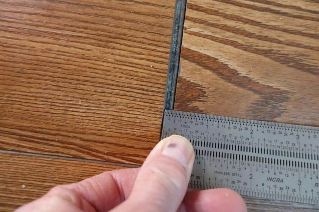 A close-up image of a wooden floor showing a gap between two floorboards. A hand is holding a ruler to measure the width of the gap, highlighting an issue with the flooring installation or wear. This is part of an inspection for potential floor refinishing by Mr Sander®.