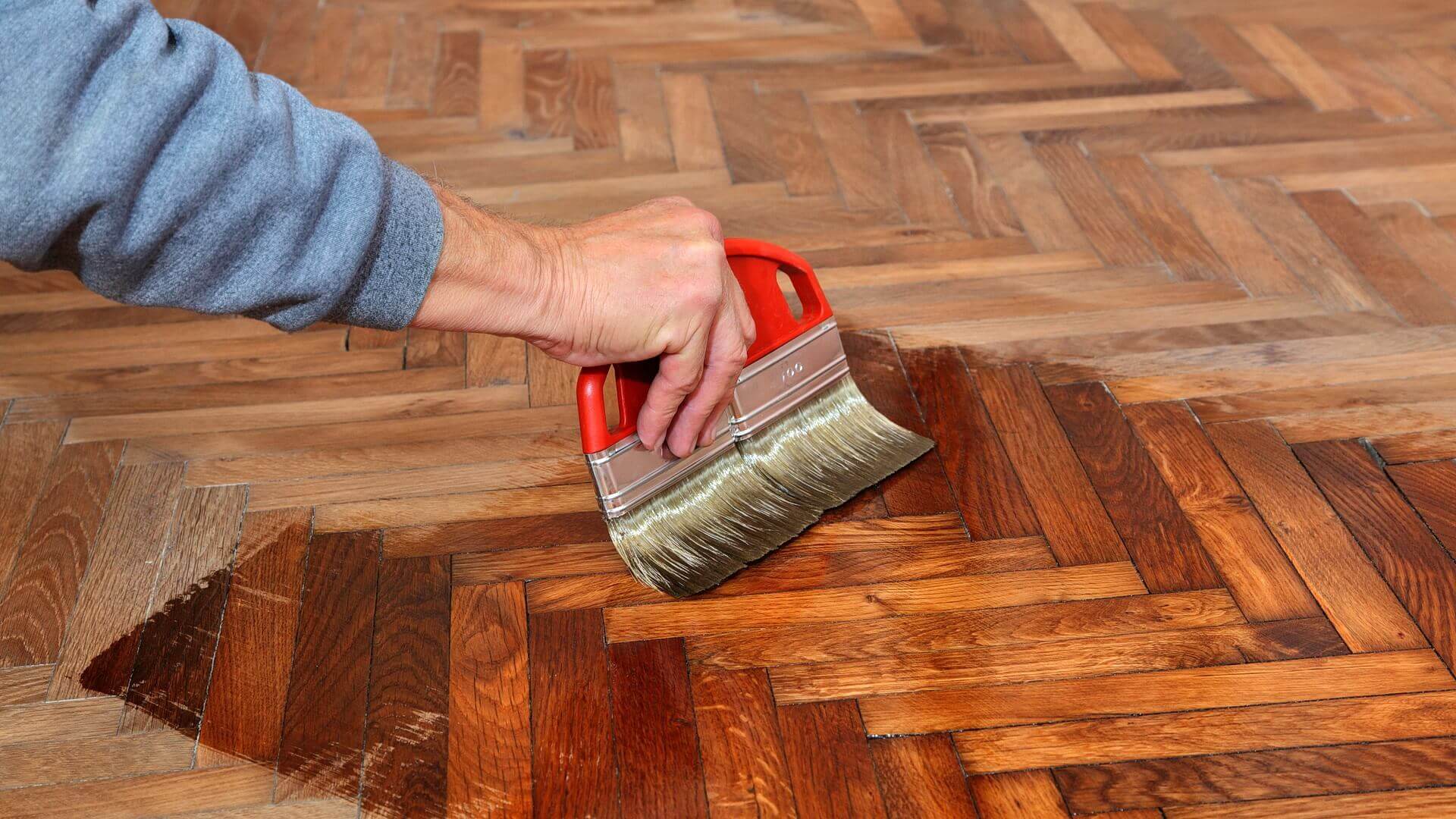 A close-up of a person using a wide brush with a red handle to apply a dark wood stain to a herringbone-patterned wooden floor. The contrast between the stained and unstained sections of the floor highlights the rich, warm tones brought out by the stain.