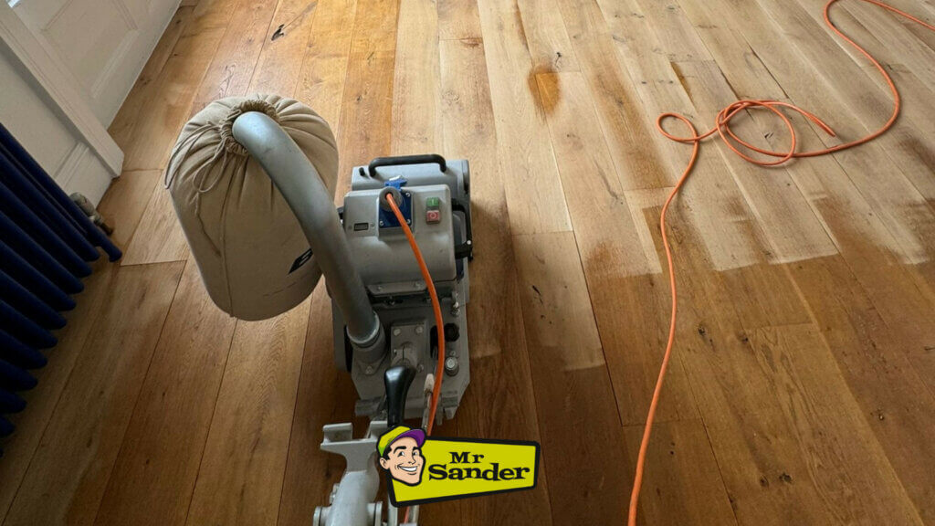 A professional from Mr Sander® is sanding a solid oak hardwood in a living room in Westcombe Park, UK using a Bona Belt sander (AMO110000.3). The machine features a 2.2 kW motor, 230 V voltage, 50 Hz frequency, and a sanding belt size of 200 x 750 mm or 250 x 750 mm. The total weight is 74 kg.
