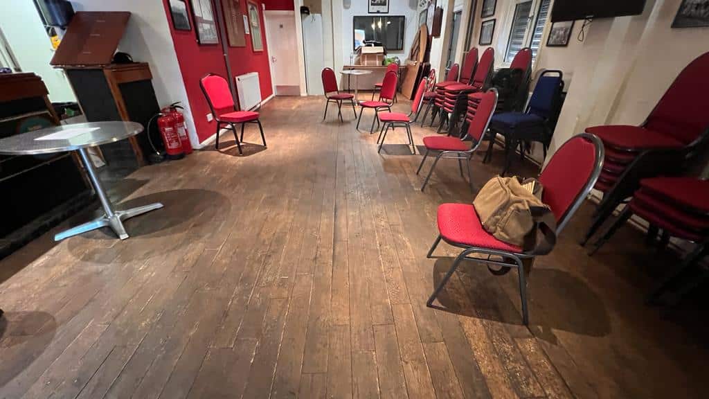 Community hall with worn, dark wooden floors in need of refinishing. The room is set up with red chairs, a few tables, and has a cozy, rustic ambiance.