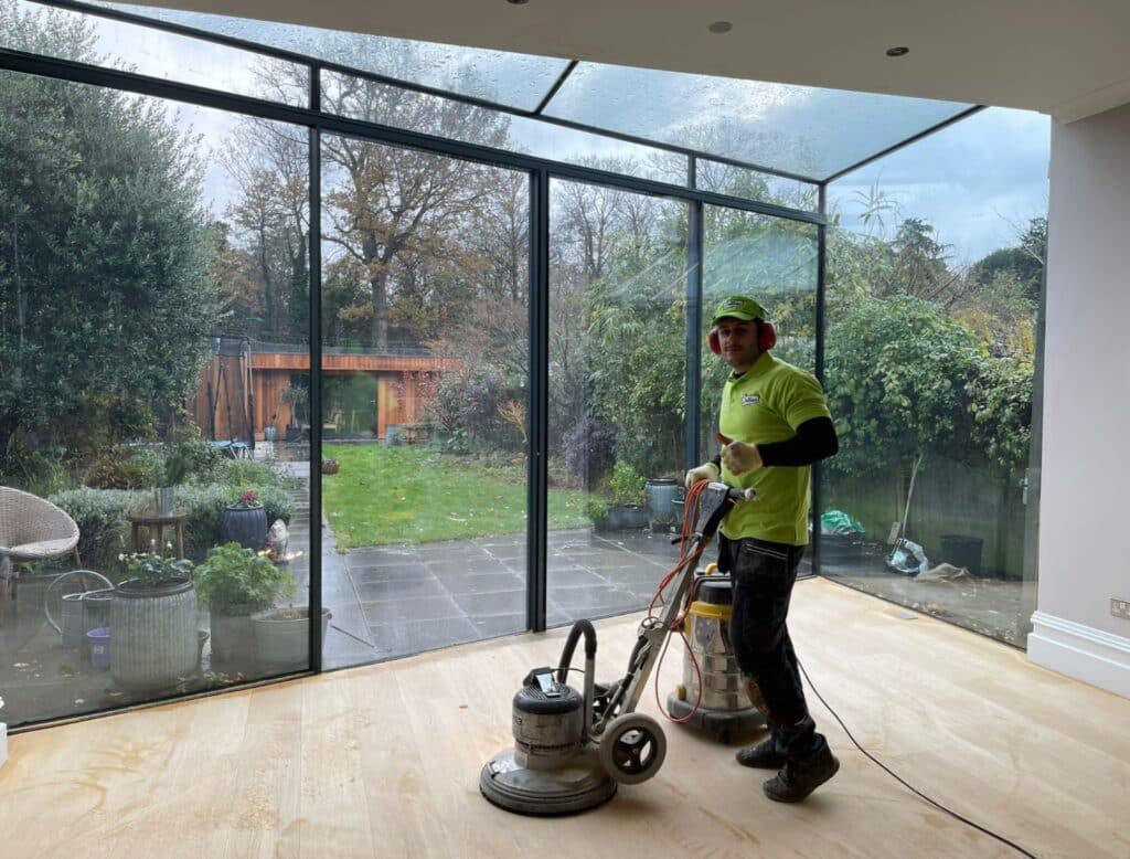 A person in bright lime green shirt, black pants, and a matching green cap stands holding a floor sander in a spacious room with a large panoramic window overlooking a garden. The garden is lush with a variety of plants, shrubs, and trees, and features a wooden structure in the background, possibly a shed or studio. There is a glass ceiling above, and it's a cloudy day outside. The hardwood floor inside shows signs of being freshly sanded, with the sander connected to an industrial vacuum to minimize dust. The person is looking towards the camera with a slight smile, appearing proud of the work being done. The interior is bright and airy, illuminated by the natural light from the expansive windows.