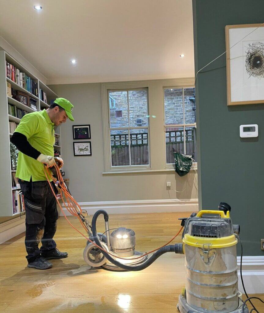 A professional from Mr Sander® sanding a hardwood floor in a De Beauvoir Town. The worker uses advanced sanding equipment with LED lighting and a dust extraction system, ensuring a smooth, clean finish. The room features modern decor with built-in bookshelves and large windows.