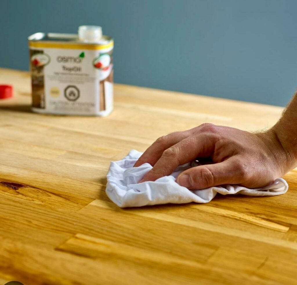 A close-up of a hand wiping a wooden countertop with a white cloth, with a can of Osmo TopOil in the background.