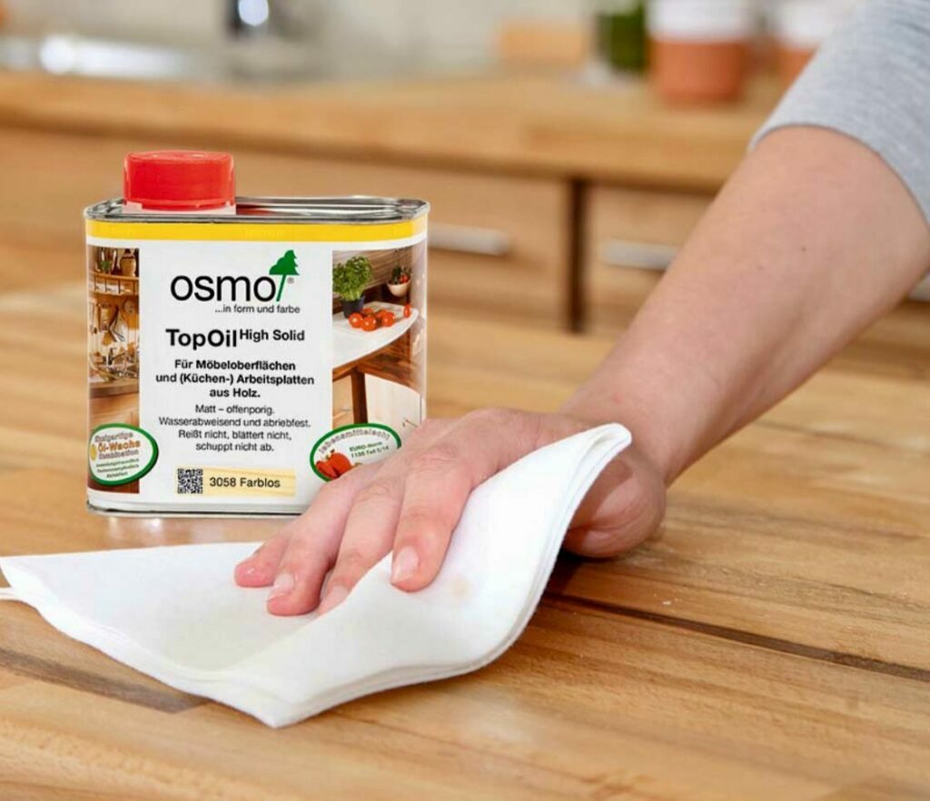 A person's hand applying a protective oil finish to a wooden surface with a cloth, next to a can of Osmo TopOil.