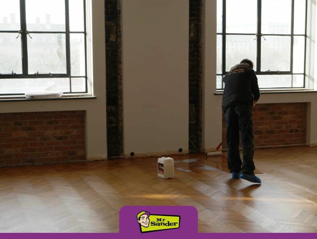 Person applying finish to a hardwood floor in a room with large windows and brick walls.