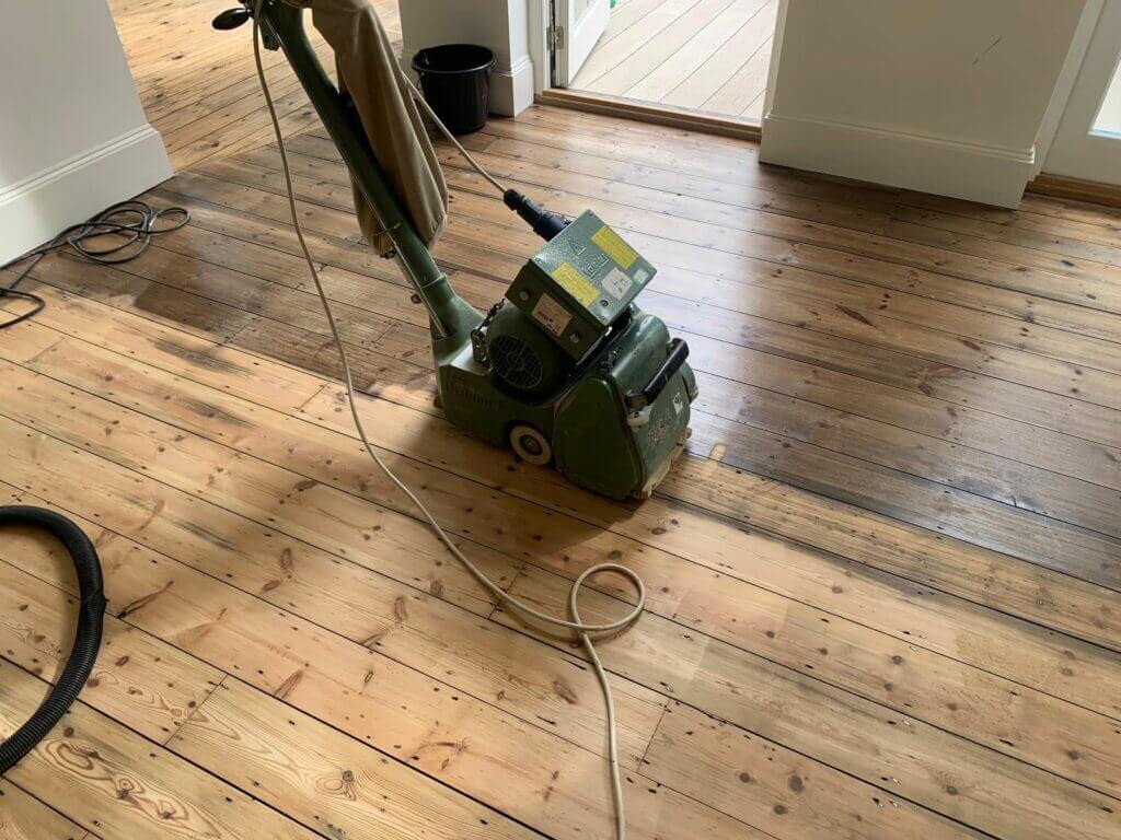 A Mr Sander® technician sands a pine floor in a living room in Elsted, UK, using a Lagler HUMMEL® floor sander. The machine features a powerful LED lighting system, high powered aggressive sanding, low dust emissions, quick belt changes, and a 230V AC motor with thermal overload protection.